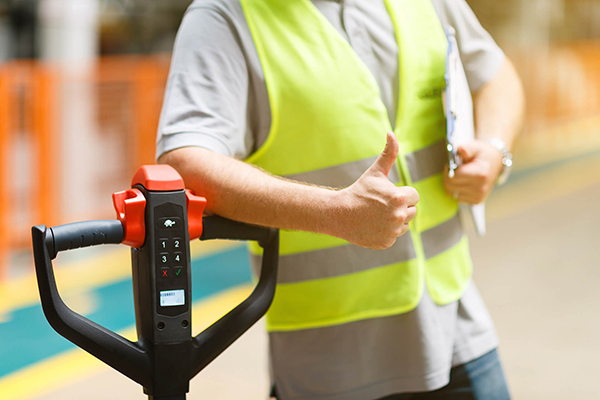 Worker leaning against pallet jack with a clipboard and thumbs up.